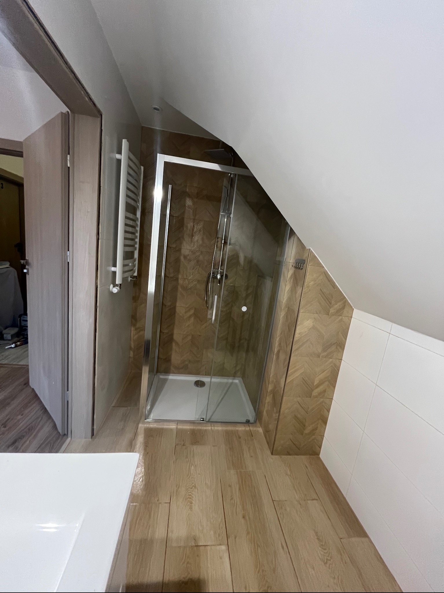 Nowoczesna łazienka na poddaszu ze szklaną kabiną prysznicową, brązowymi płytkami w jodełkę i białymi kaflami, widok na grzejnik i fragment blatu umywalki.