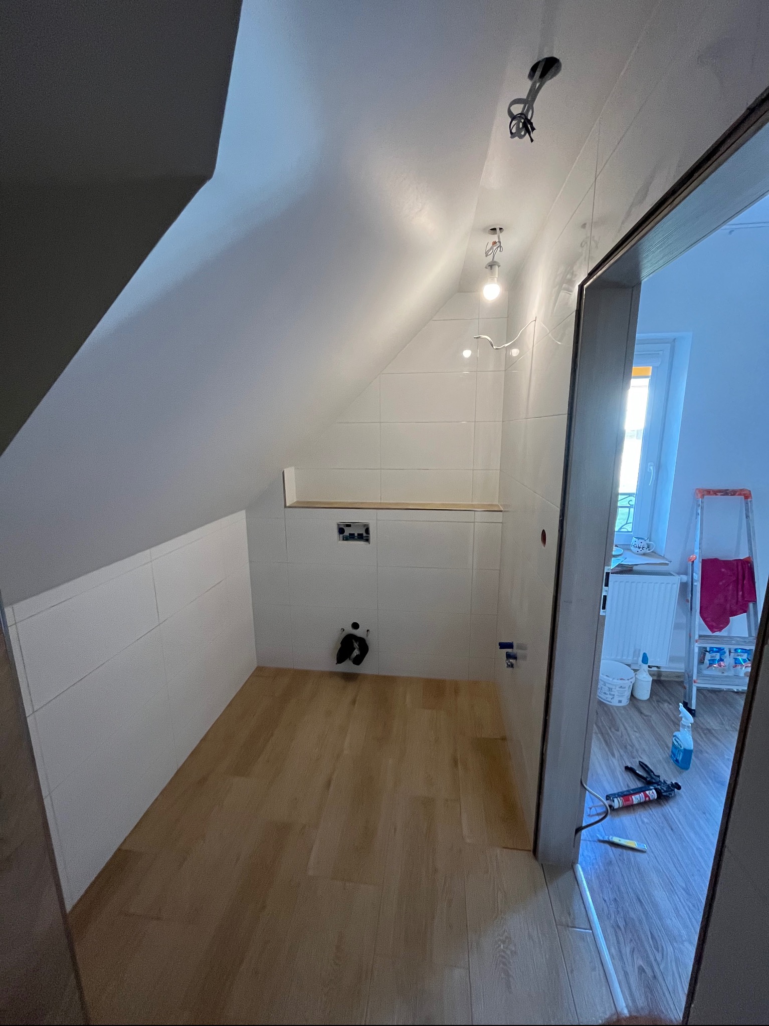 Remontowana łazienka na poddaszu: białe płytki, drewniana podłoga, instalacja oświetleniowa w suficie, widoczne narzędzia i drabina w tle.