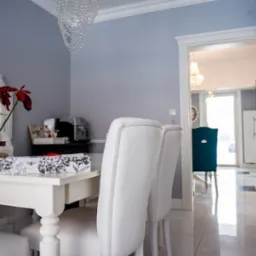 Jasne, przestronne wnętrze jadalni z białym stołem, krzesłami obitymi skórą, kwiatem poinsecji w doniczce i kryształowym żyrandolem, widoczne przez otwarte drzwi kolejne pomieszczenie.