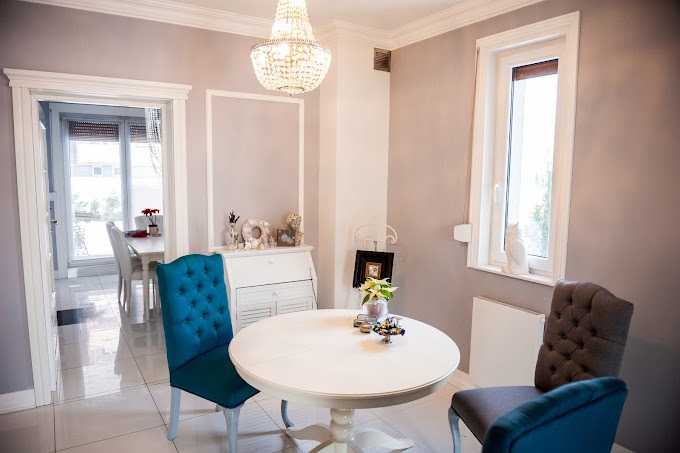 Jasne wnętrze jadalni z okrągłym białym stołem, krzesłami z turkusową tapicerką, kryształowym żyrandolem i dekoracjami w stylu vintage.