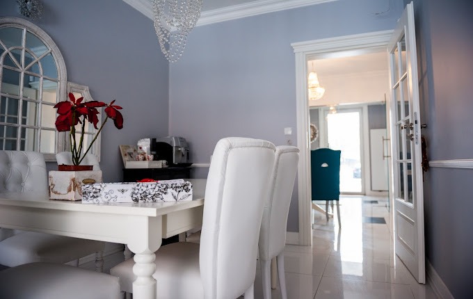 Jasne, przestronne wnętrze jadalni z białym stołem, krzesłami obitymi skórą, kwiatem poinsecji w doniczce i kryształowym żyrandolem, widoczne przez otwarte drzwi kolejne pomieszczenie.