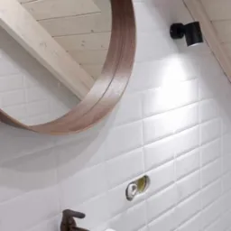 Nowoczesna łazienka na poddaszu z białą umywalką, brązową armaturą i okrągłym lustrem z drewnianą ramą, oświetlona czarnym reflektorem na tle białych, prostokątnych płytek.