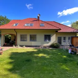 Dom jednorodzinny z czerwonym dachem, elewacja częściowo przygotowana do prac, okna zabezpieczone folią, widoczne narzędzia i drabina, trawnik przed domem.