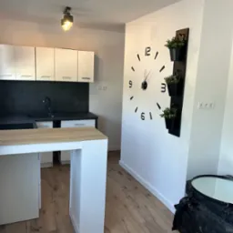 Nowoczesna kuchnia z białymi szafkami, czarnym blatem i dużą ścianą z zegarem oraz pionowym kwietnikiem, drewniana podłoga w trakcie prac wykończeniowych.