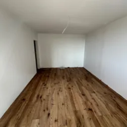 Pokój po ułożeniu drewnopodobnych paneli podłogowych, widoczne białe ściany i sufit, wystające kable ze ściany i listwy dystansowe przy ścianach.