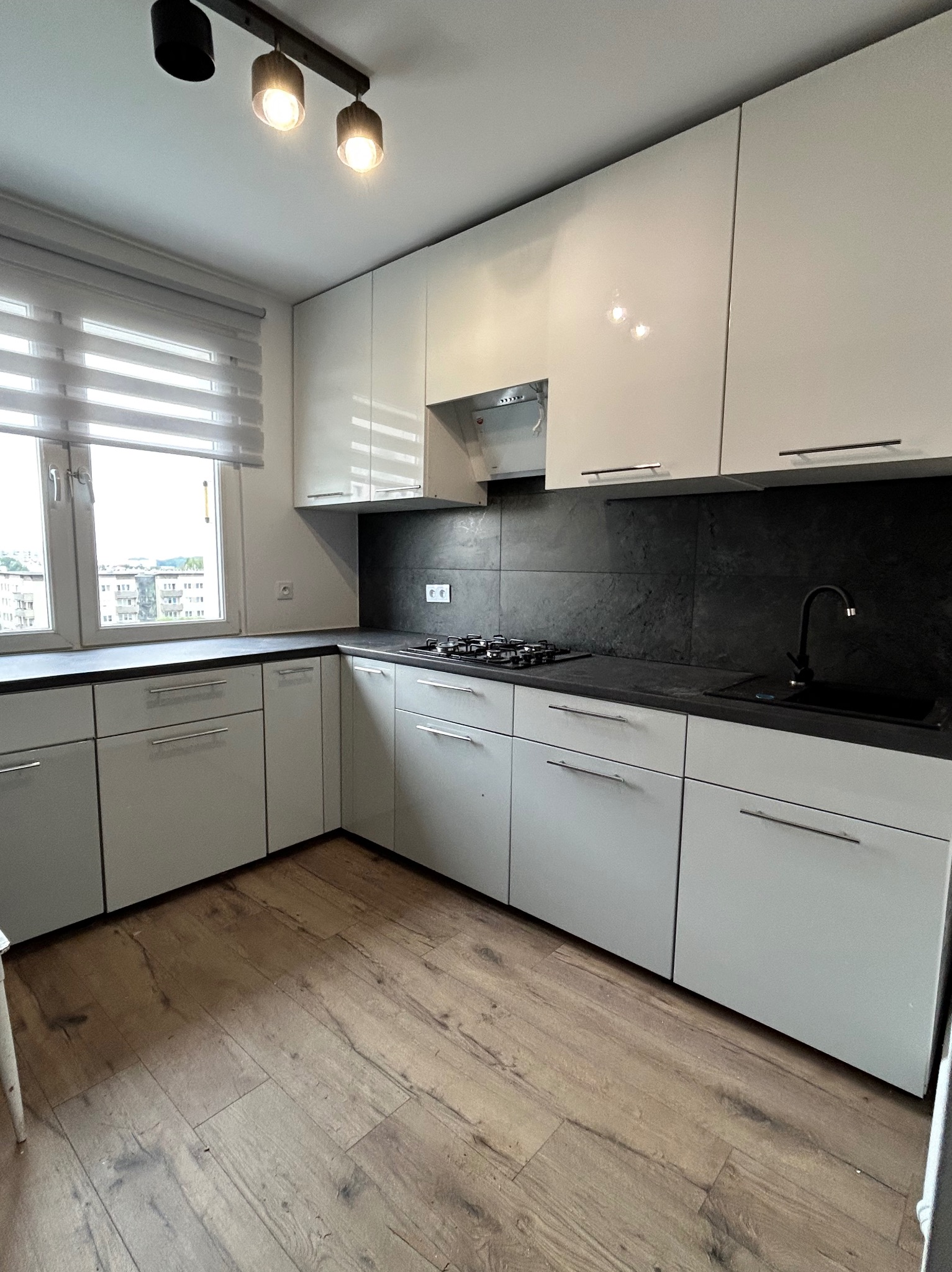 Nowoczesna kuchnia z białymi, błyszczącymi szafkami, czarnym blatem i panelami podłogowymi w odcieniach brązu. Widoczne okno z żaluzjami i oświetlenie sufitowe.