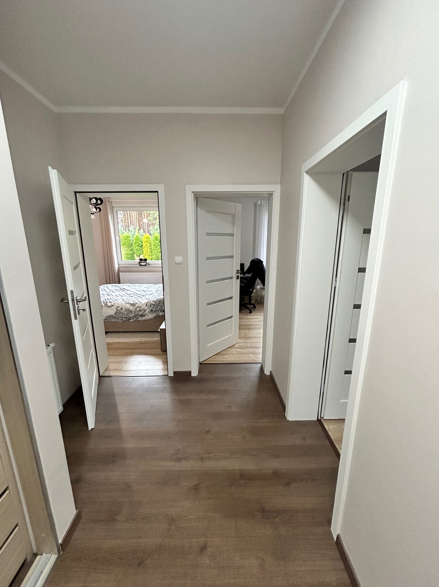 Korytarz z trzema otwartymi drzwiami, widok na pokój z łóżkiem i biuro, panele podłogowe w odcieniach brązu, ściany pomalowane na jasny kolor.