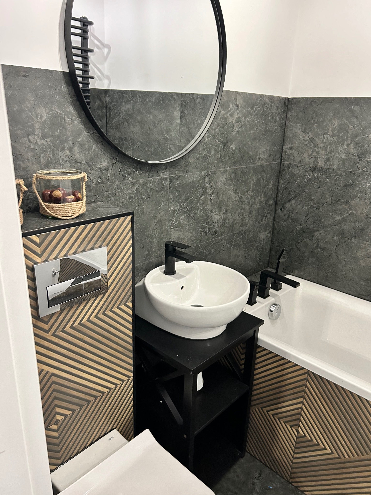 Nowoczesna łazienka z okrągłym lustrem, umywalką nablatową na czarnym regale, szarą kamienną okładziną ścienną oraz drewnianą mozaiką na zabudowie toalety i obudowie wanny.