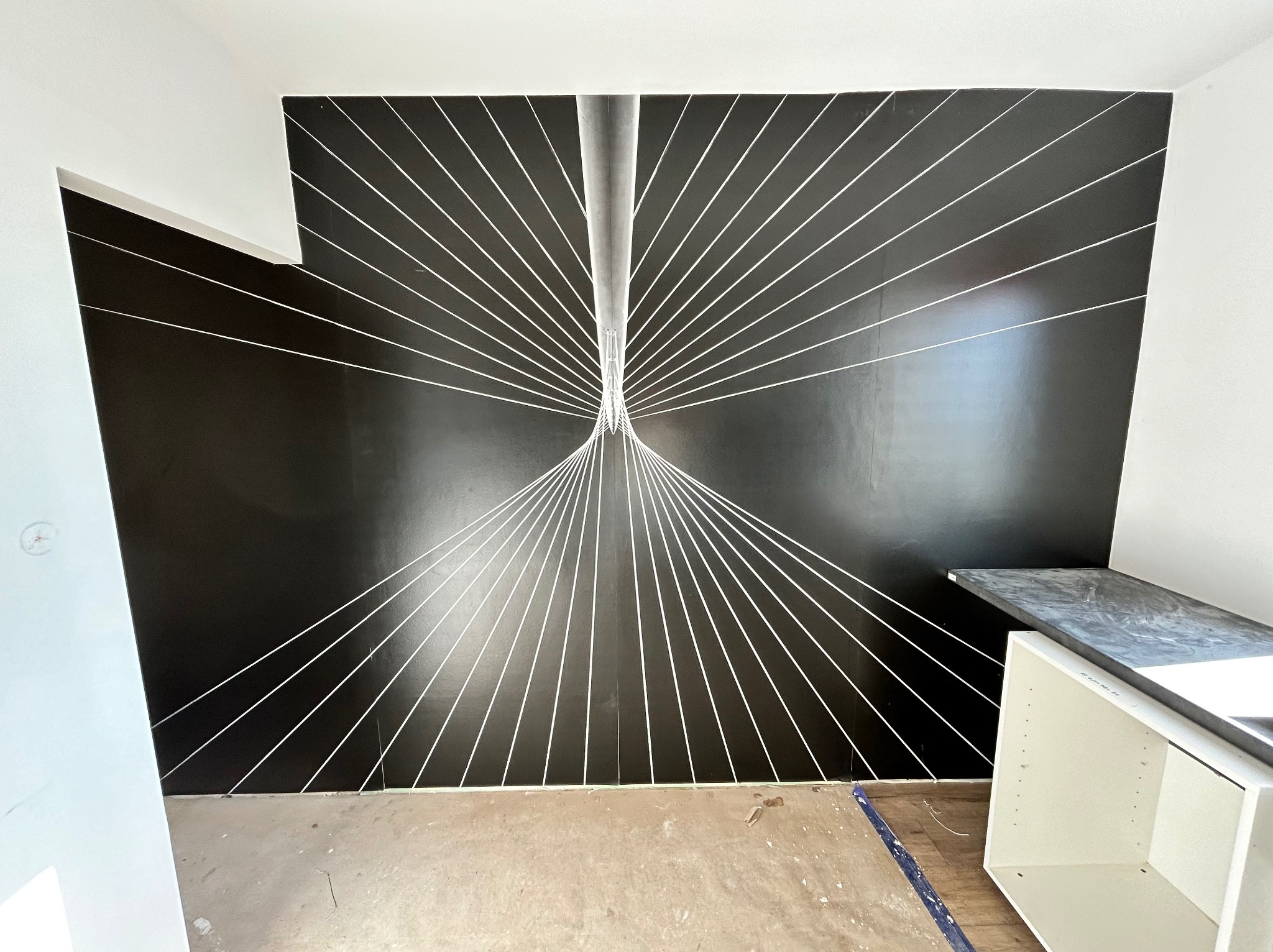 Czarna ściana z geometrycznym wzorem białych linii, przypominającym perspektywę mostu linowego, w trakcie prac wykończeniowych, widoczny fragment niedokończonej podłogi i szafka kuchenna bez frontów.