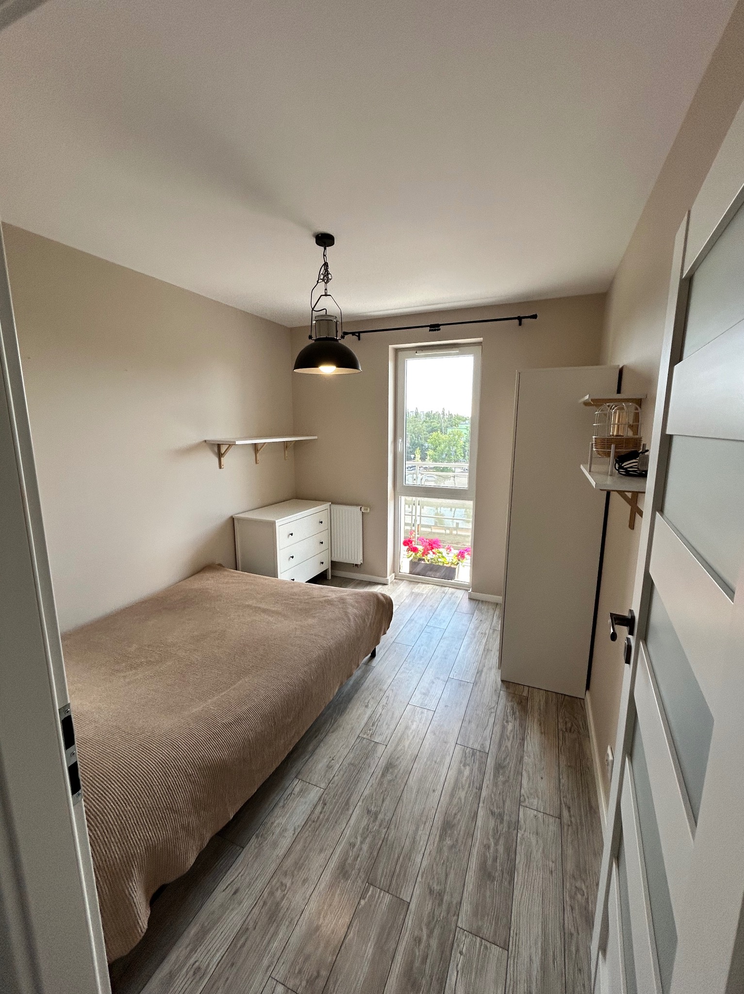 Jasne wnętrze pokoju z łóżkiem, komodą, półkami i podłogą z paneli, widok przez otwarte drzwi na balkon z kwiatami.