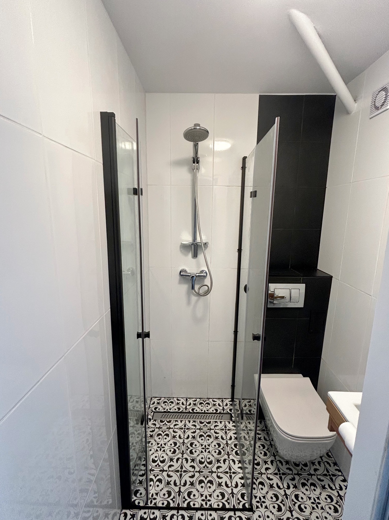 Nowoczesna łazienka z kabiną prysznicową typu walk-in z czarnymi ramami, białą armaturą i czarno-białą mozaiką podłogową, widoczne płytki ścienne w dwóch kolorach: białym i czarnym.