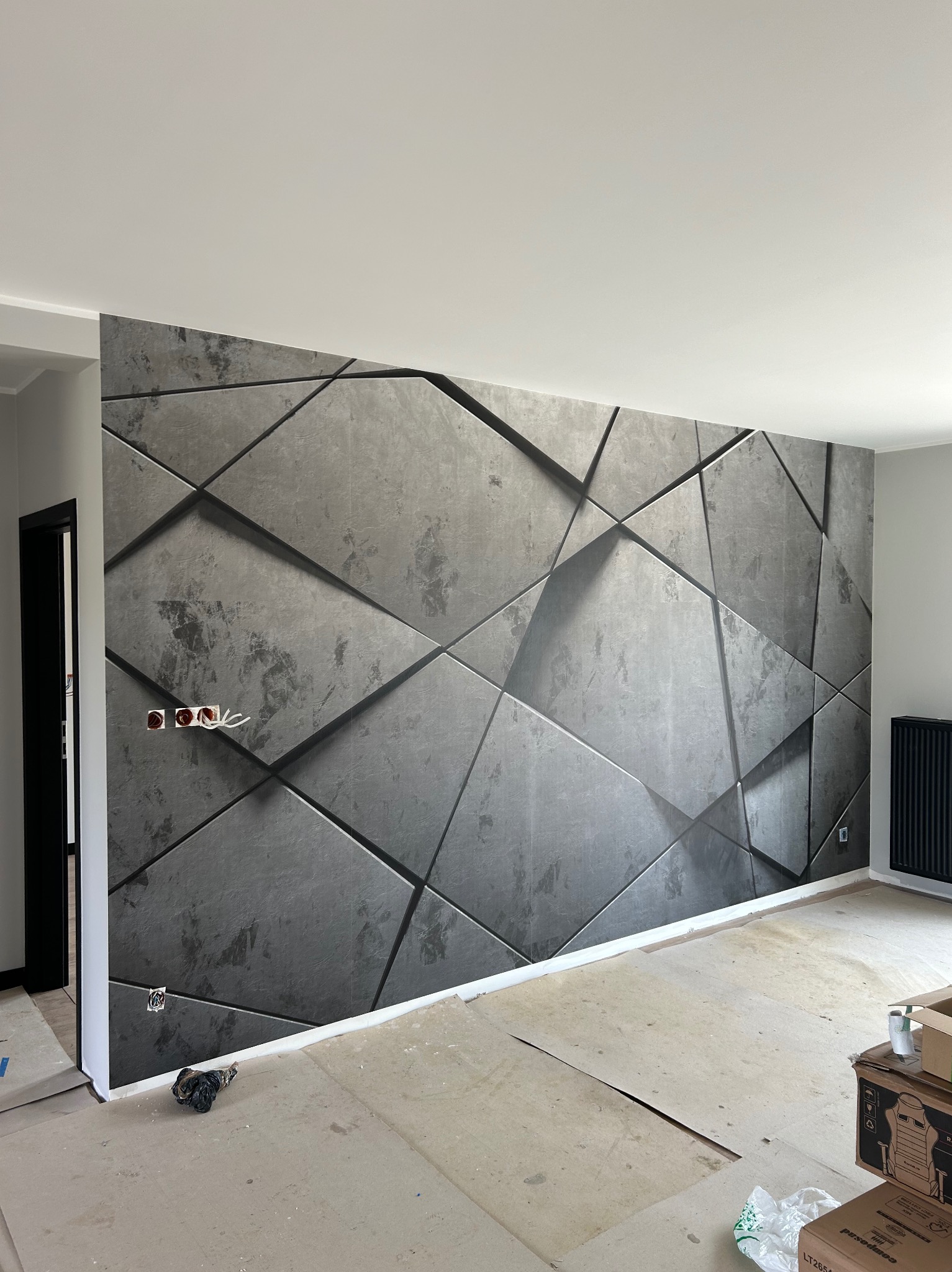 Pokój w trakcie wykańczania z fototapetą o geometrycznym wzorze imitującym betonowe płyty 3D, widoczne puszki elektryczne bez osprzętu i prowizoryczna podłoga.