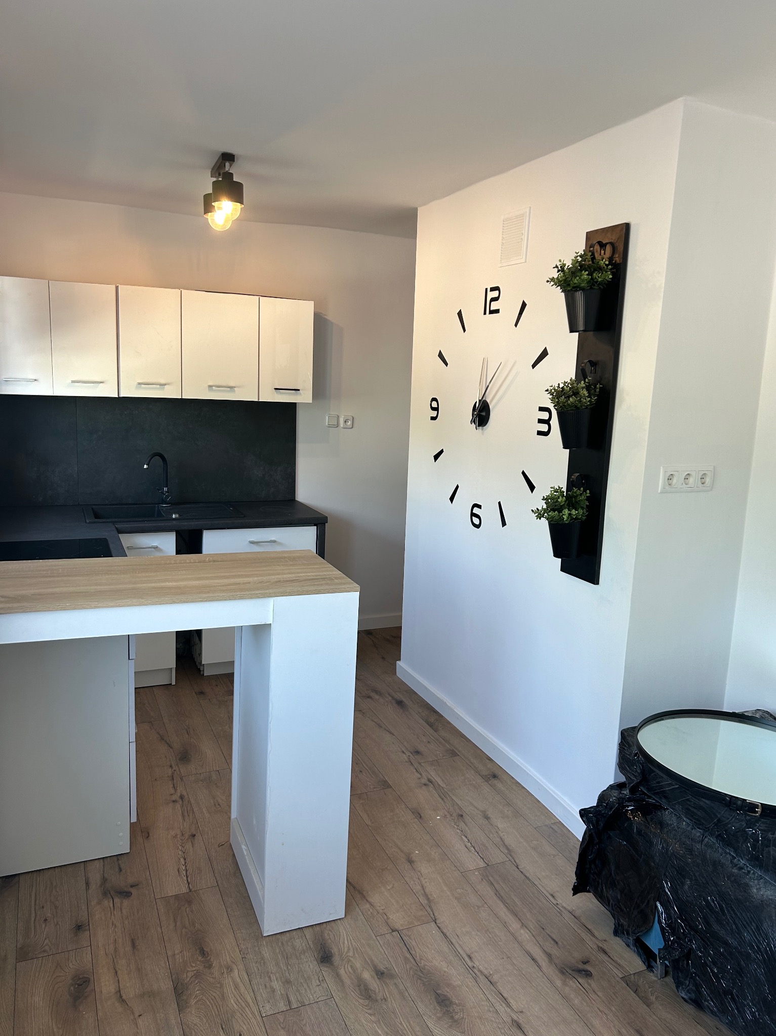 Nowoczesna kuchnia z białymi szafkami, czarnym blatem i dużą ścianą z zegarem oraz pionowym kwietnikiem, drewniana podłoga w trakcie prac wykończeniowych.