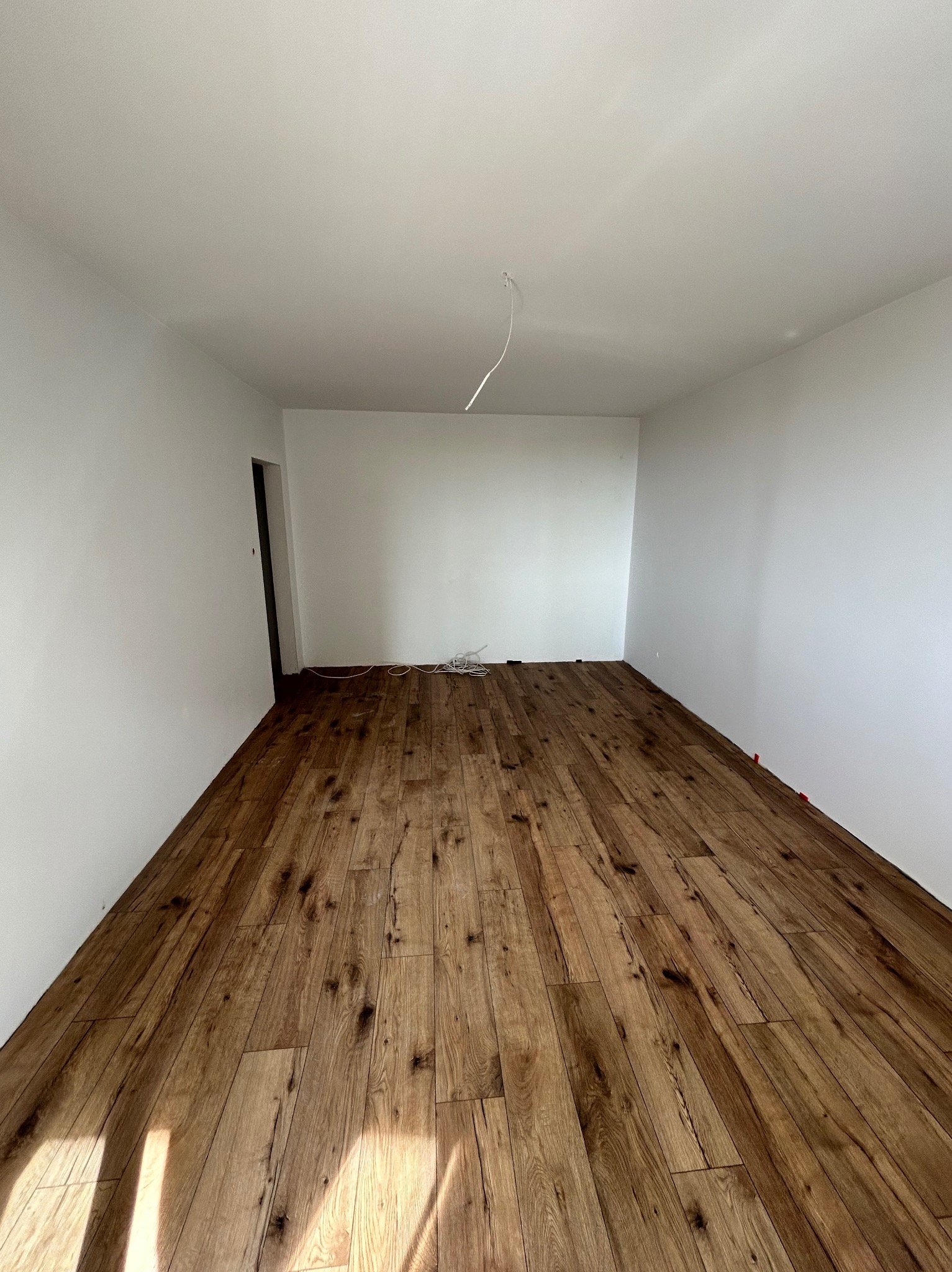 Pokój po ułożeniu drewnopodobnych paneli podłogowych, widoczne białe ściany i sufit, wystające kable ze ściany i listwy dystansowe przy ścianach.