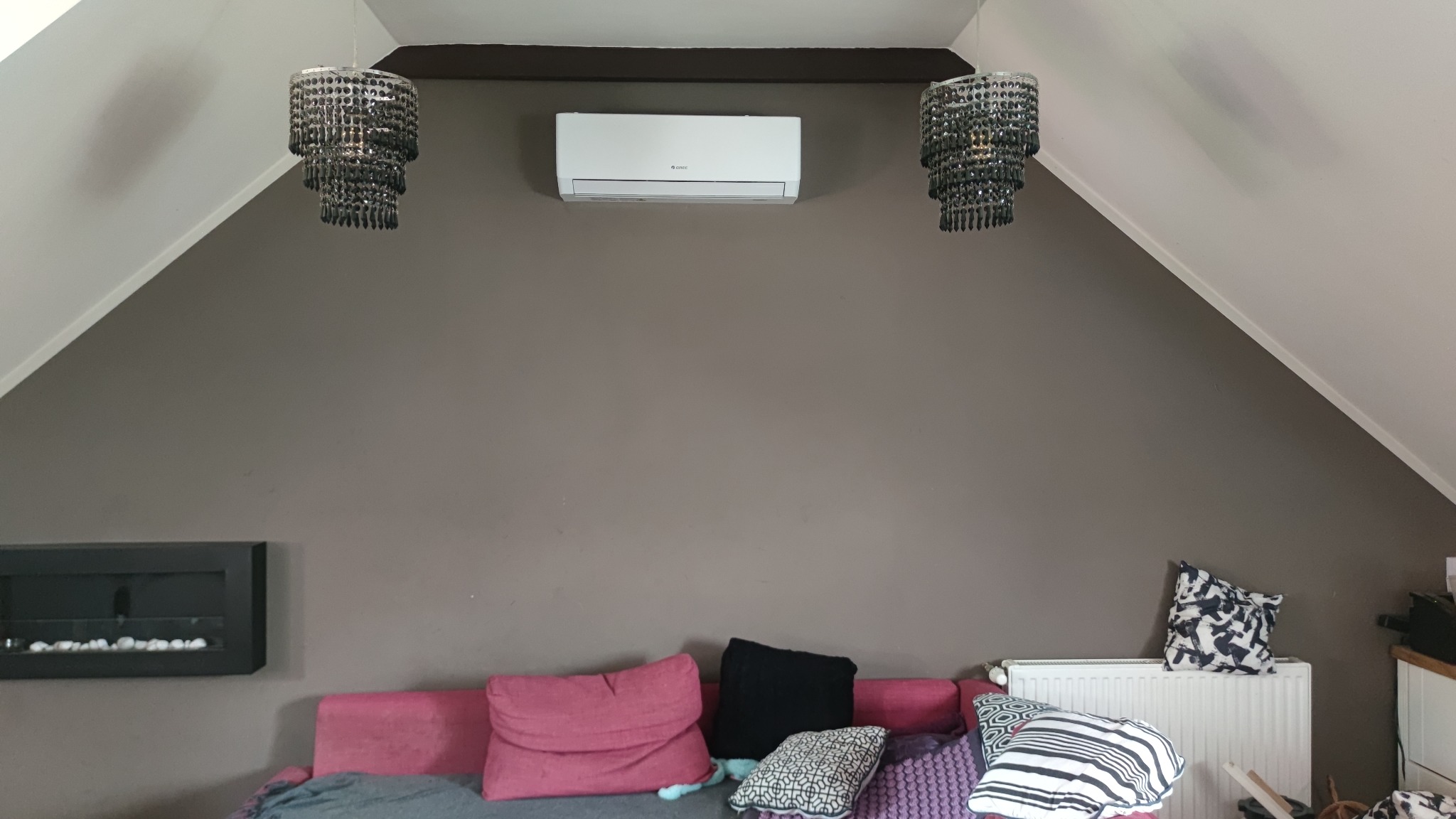 Klimatyzator Gree zamontowany na szarej, skośnej ścianie w pokoju na poddaszu, pomiędzy dwoma ozdobnymi, czarnymi żyrandolami, nad różową sofą z poduszkami.
