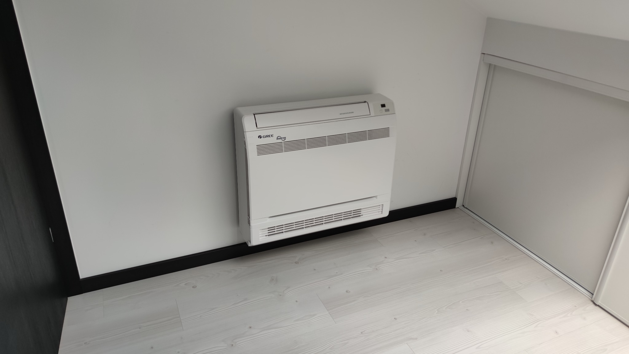 Biała, przypodłogowa jednostka klimatyzacyjna marki Gree zamontowana w rogu jasnego pokoju z panelami podłogowymi i przesuwnymi drzwiami szafy.