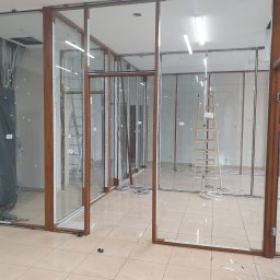 Modern Glass Wojciech Śnieżek - Montaż szklanych ścianek działowych z drewnianymi ramami. Widoczne drabiny, kable i elementy montażowe. Prace w toku, widok na wnętrze pomieszczenia.