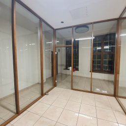 Modern Glass Wojciech Śnieżek - Szklana zabudowa biurowa z drewnianymi ramami, widok na kafelkowaną podłogę i okna w tle, jasne wnętrze, widoczne gniazdka elektryczne na ścianie.