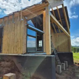 Surowy stan domu z drewnianą konstrukcją szkieletową, widoczne okno i elementy dachu, w tle błękitne niebo z chmurami, teren budowy z ziemią i zielenią.