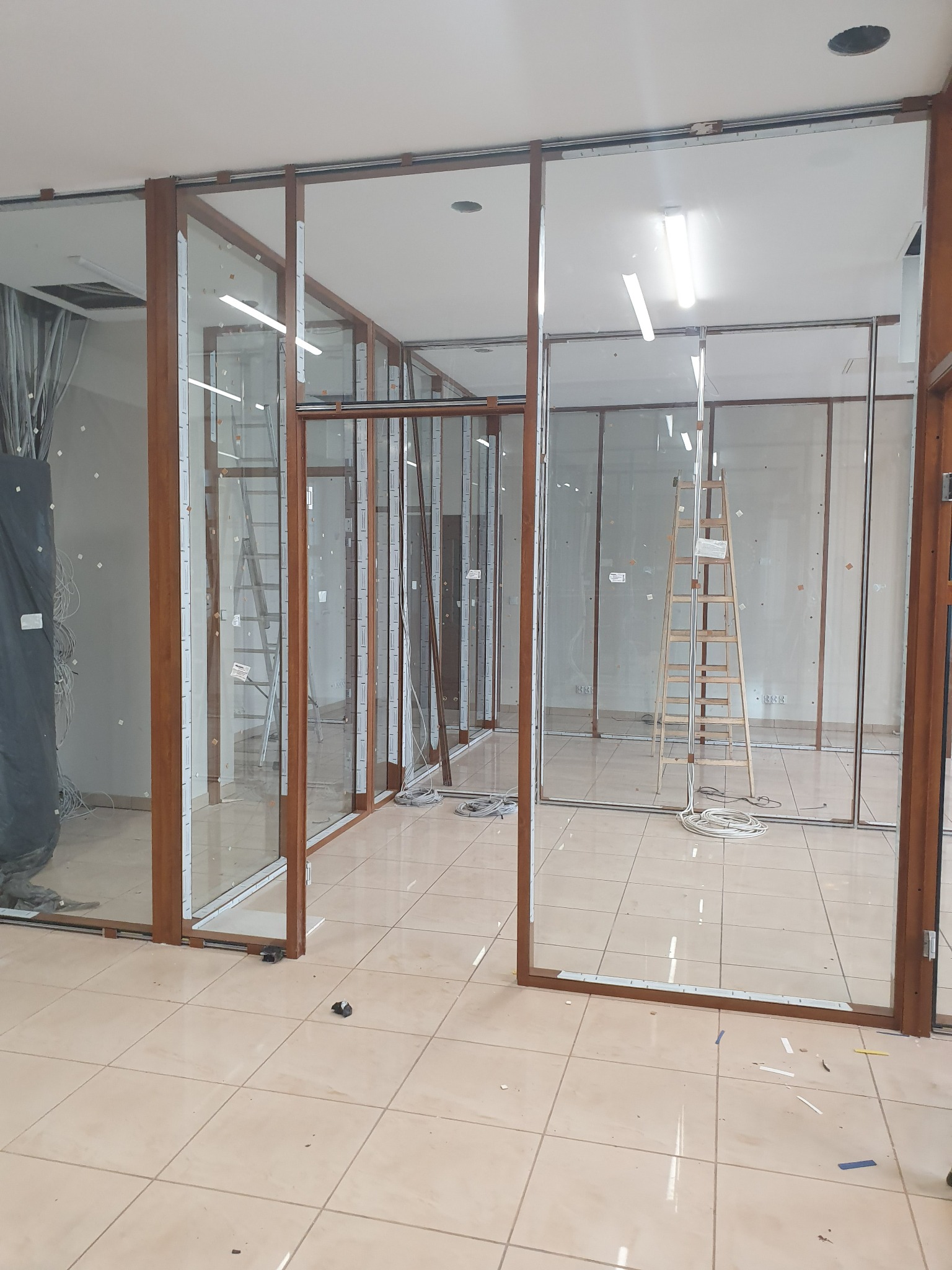 Montaż szklanych ścianek działowych z drewnianymi ramami. Widoczne drabiny, kable i elementy montażowe. Prace w toku, widok na wnętrze pomieszczenia.