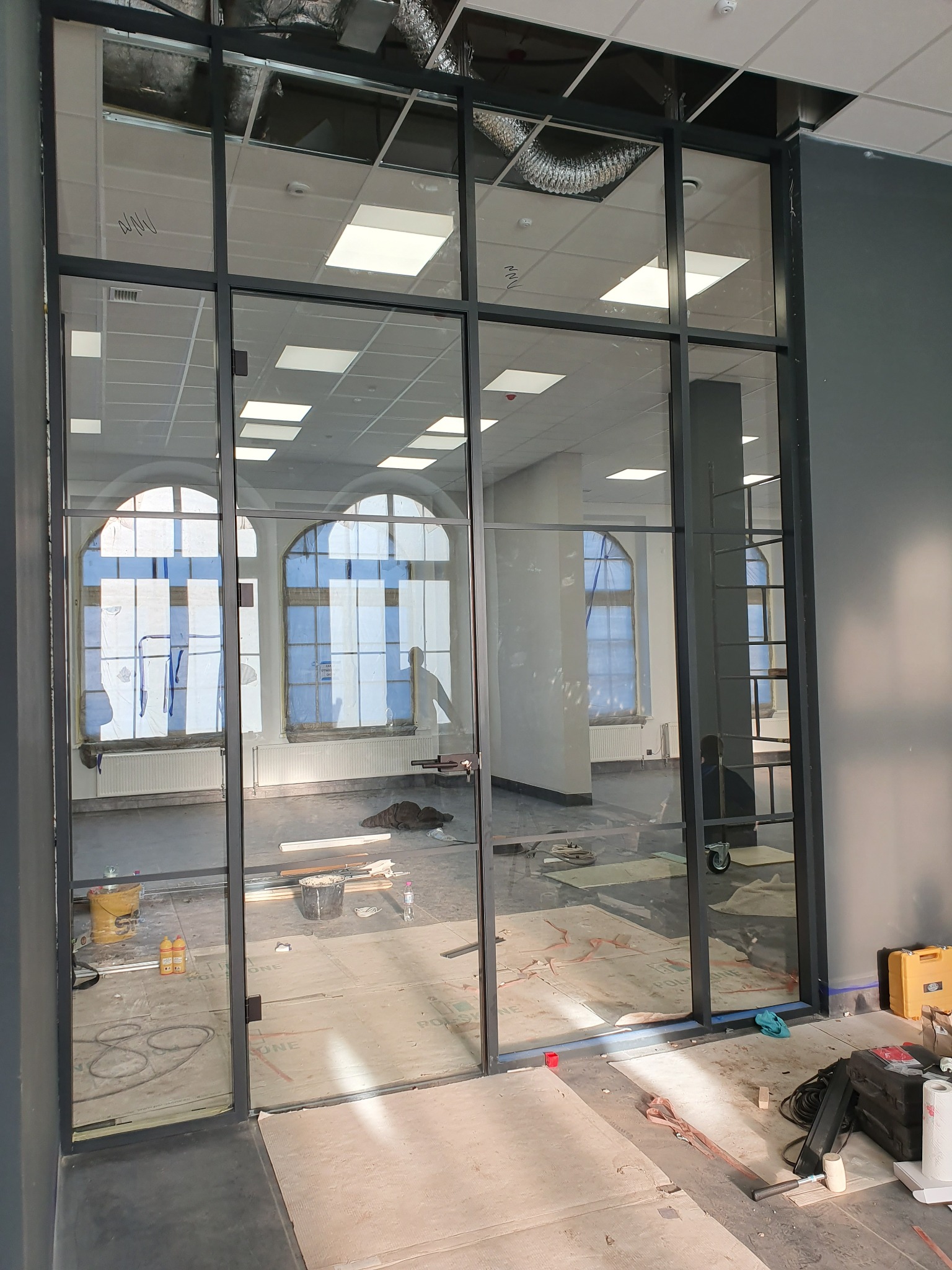 Szklana ściana działowa z czarnymi profilami, widok na wnętrze biura z dużymi oknami. Elementy montażowe i narzędzia na podłodze sugerują trwające prace.