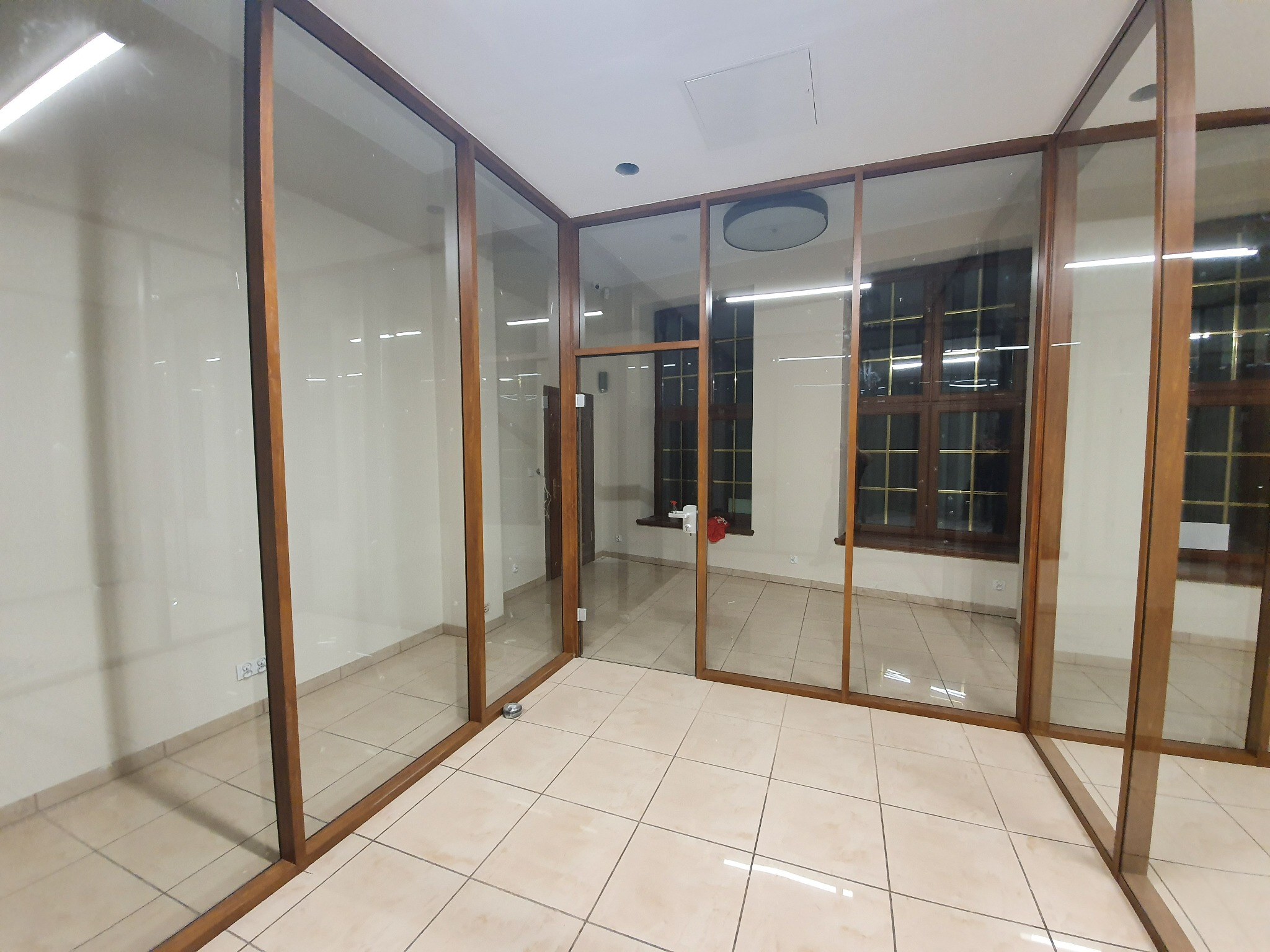 Szklana zabudowa biurowa z drewnianymi ramami, widok na kafelkowaną podłogę i okna w tle, jasne wnętrze, widoczne gniazdka elektryczne na ścianie.