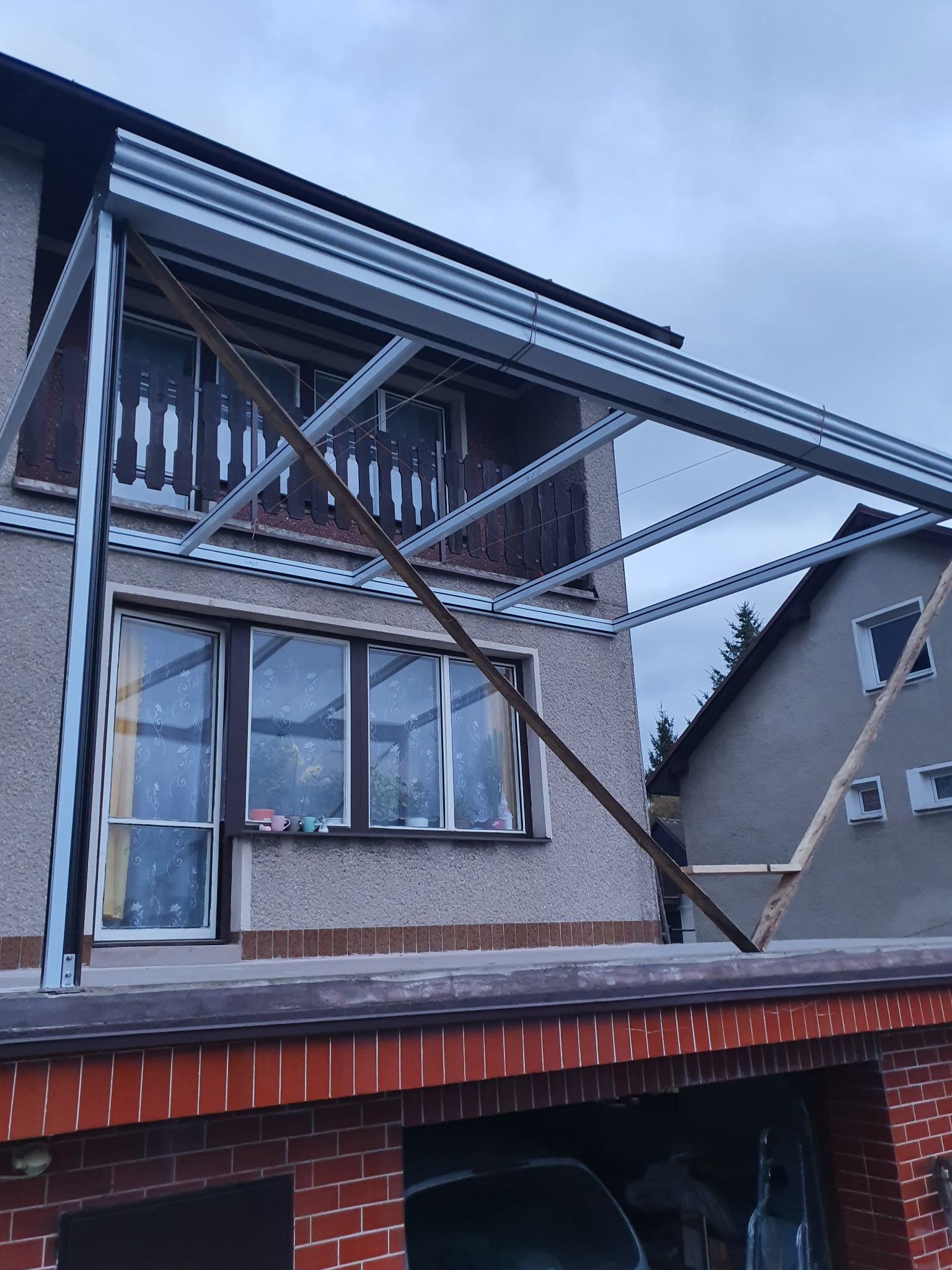Montaż zadaszenia balkonu z metalową konstrukcją, widok od dołu, wspartej na drewnianych podporach, na tle elewacji budynku i pochmurnego nieba.
