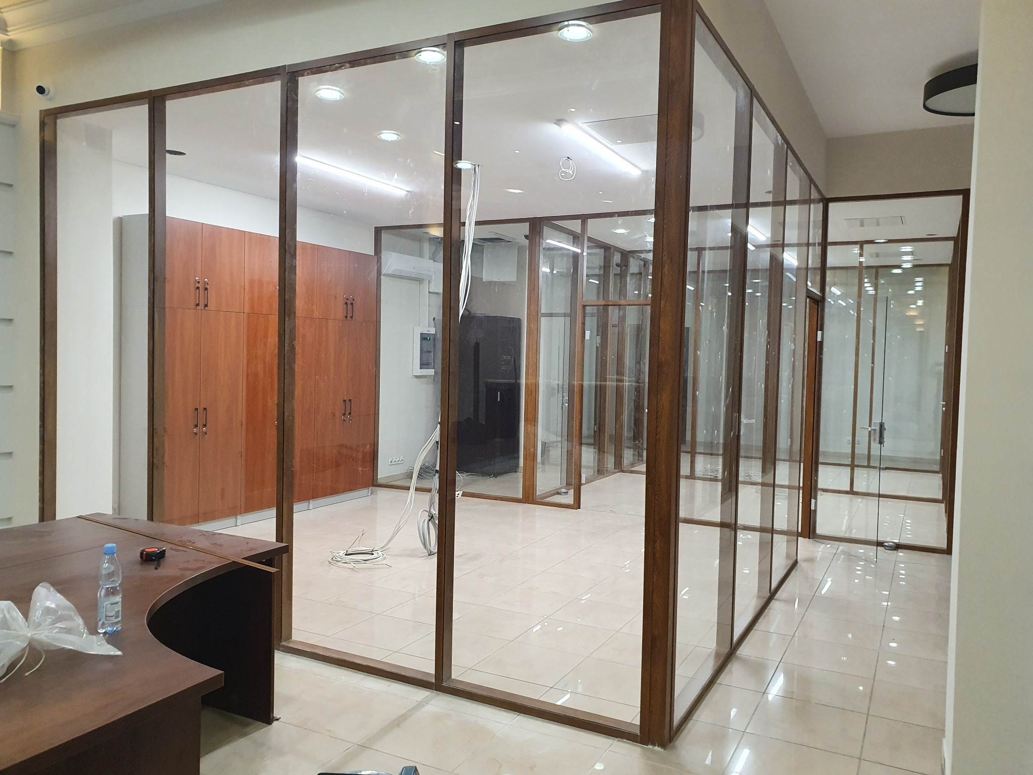 Szklane ścianki działowe z brązowymi ramami tworzą nowoczesne biuro. Widoczny fragment biurka z butelką wody i miarką. Jasne wnętrze, płytki podłogowe.