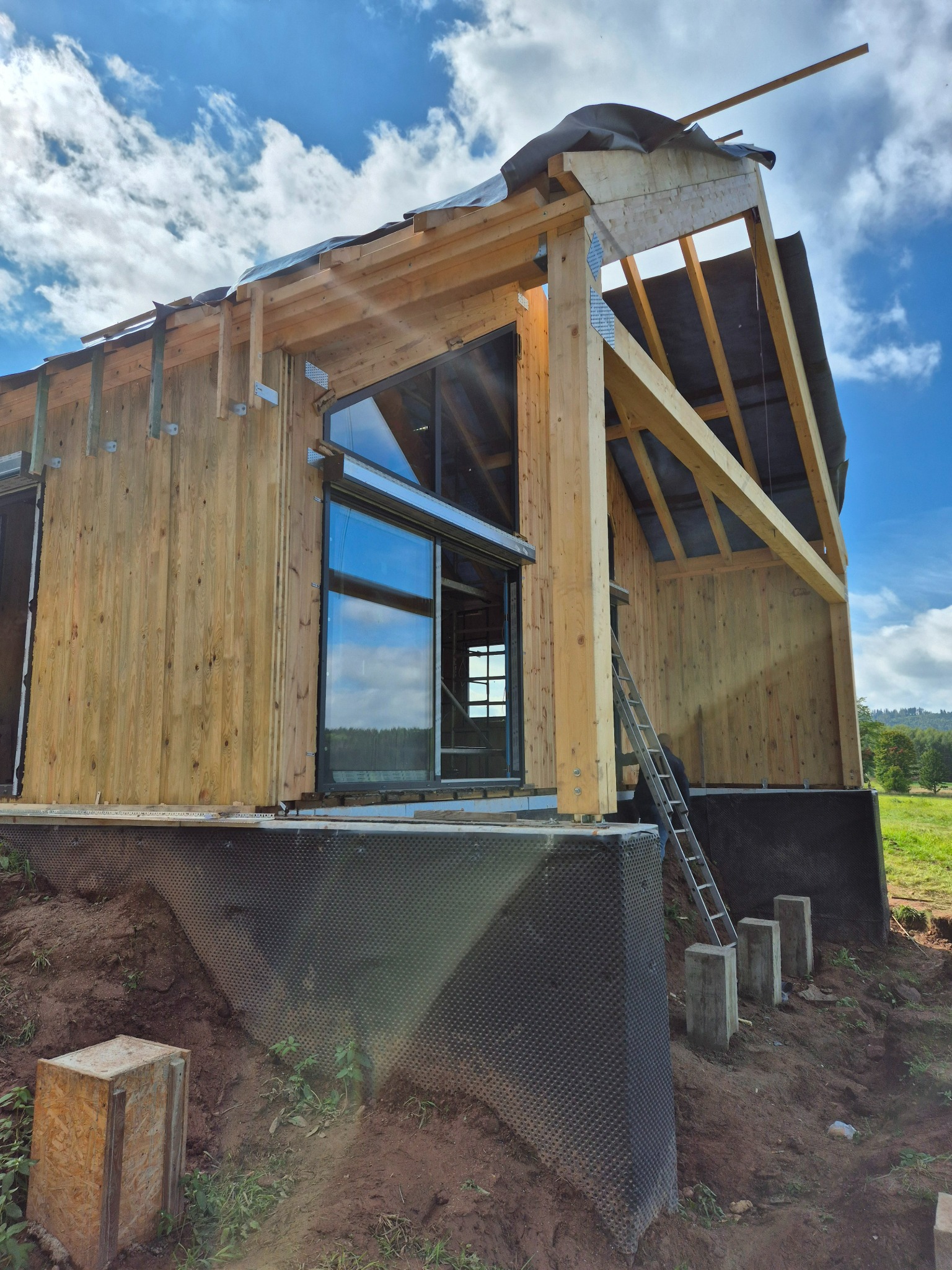 Surowy stan domu z drewnianą konstrukcją szkieletową, widoczne okno i elementy dachu, w tle błękitne niebo z chmurami, teren budowy z ziemią i zielenią.