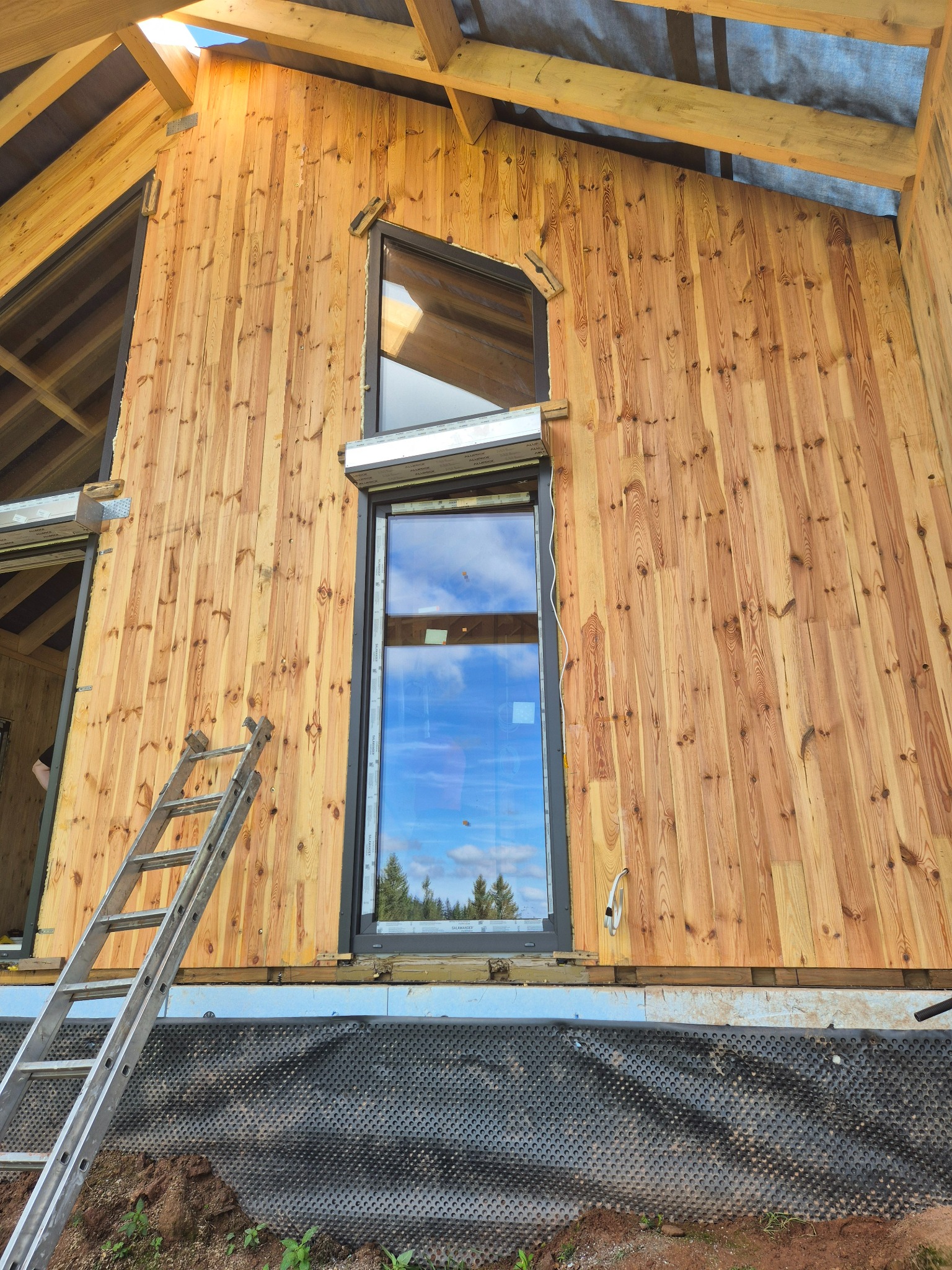 Montaż okien z roletami zewnętrznymi w drewnianym domu w trakcie budowy. Widoczna drabina oparta o ścianę i fragment czarnej folii fundamentowej.