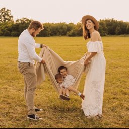 juliah.Fotografia - Rodzina z dwójką dzieci bawi się na łące, trzymając koc jak hamak, tata w trampkach, mama w ażurowej sukience i kapeluszu, ciepłe światło zachodzącego słońca.