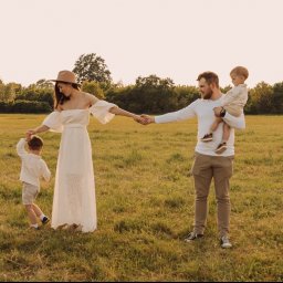 juliah.Fotografia - Sesja rodzinna we Wrocławiu: Mama w białej sukience i kapeluszu prowadzi za rękę synka, tata trzyma drugiego synka na rękach, wszyscy na łące w ciepłym świetle.