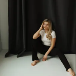Portret kobiety siedzącej na podłodze na tle czarnej zasłony, ubrana w biały t-shirt i czarne spodnie, z dłonią opartą o głowę, patrząca w obiektyw.