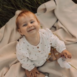 juliah.Fotografia - Uśmiechnięta, rudowłosa dziewczynka w kwiecistym ubranku siedzi na beżowym kocyku rozłożonym na trawie.