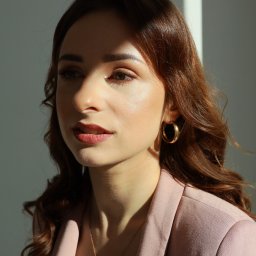 juliah.Fotografia - Portret kobiety w pudrowo-różowym żakiecie, z delikatnym makijażem i złotymi kolczykami, oświetlonej kontrastowym światłem słonecznym, z zamyślonym wyrazem twarzy.