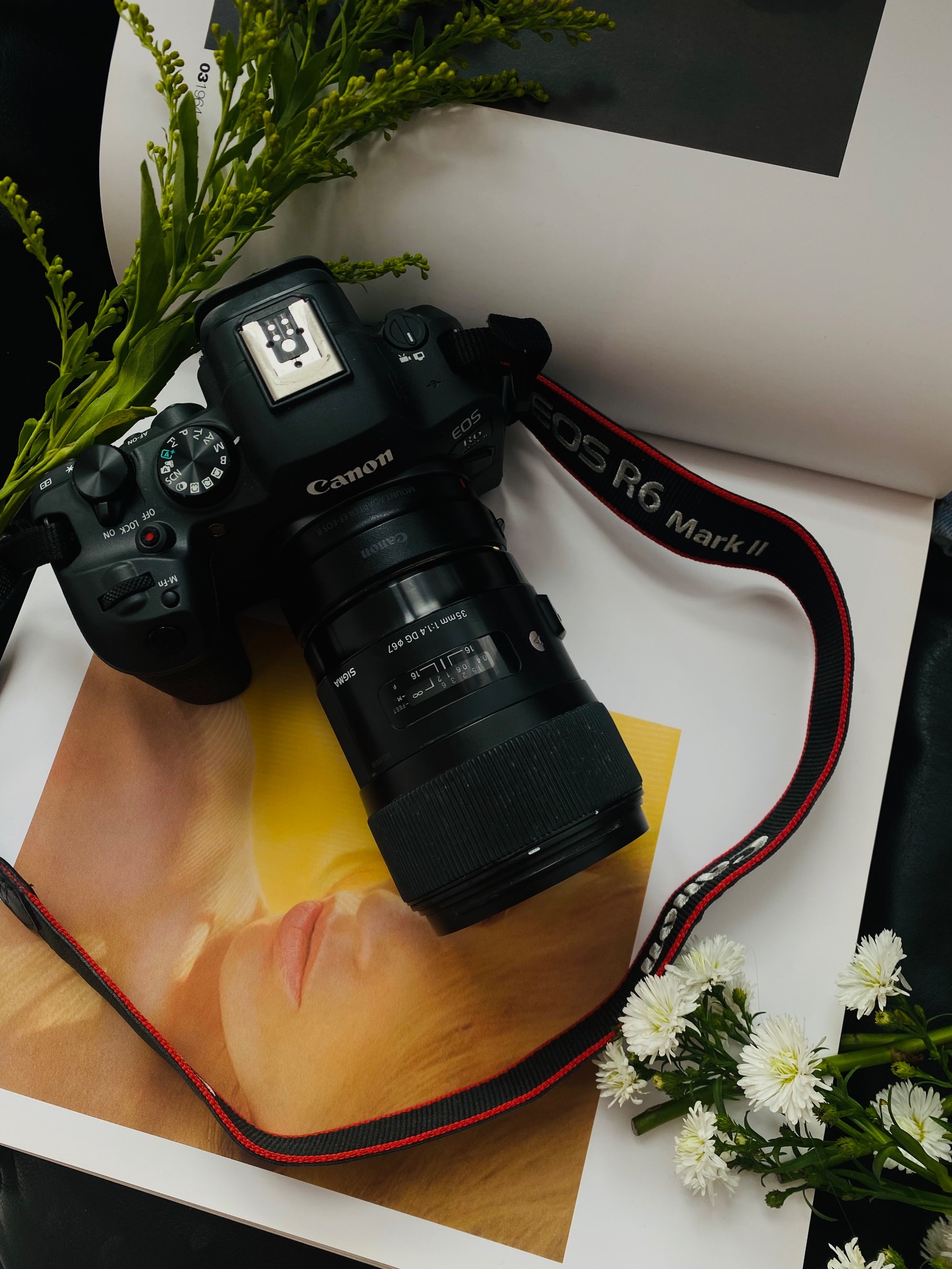 Aparat Canon EOS R6 Mark II z obiektywem Sigma 35mm f/1.4 DG, ułożony na otwartej książce z wizerunkiem twarzy modelki, ozdobiony gałązkami zieleni i białymi kwiatami.
