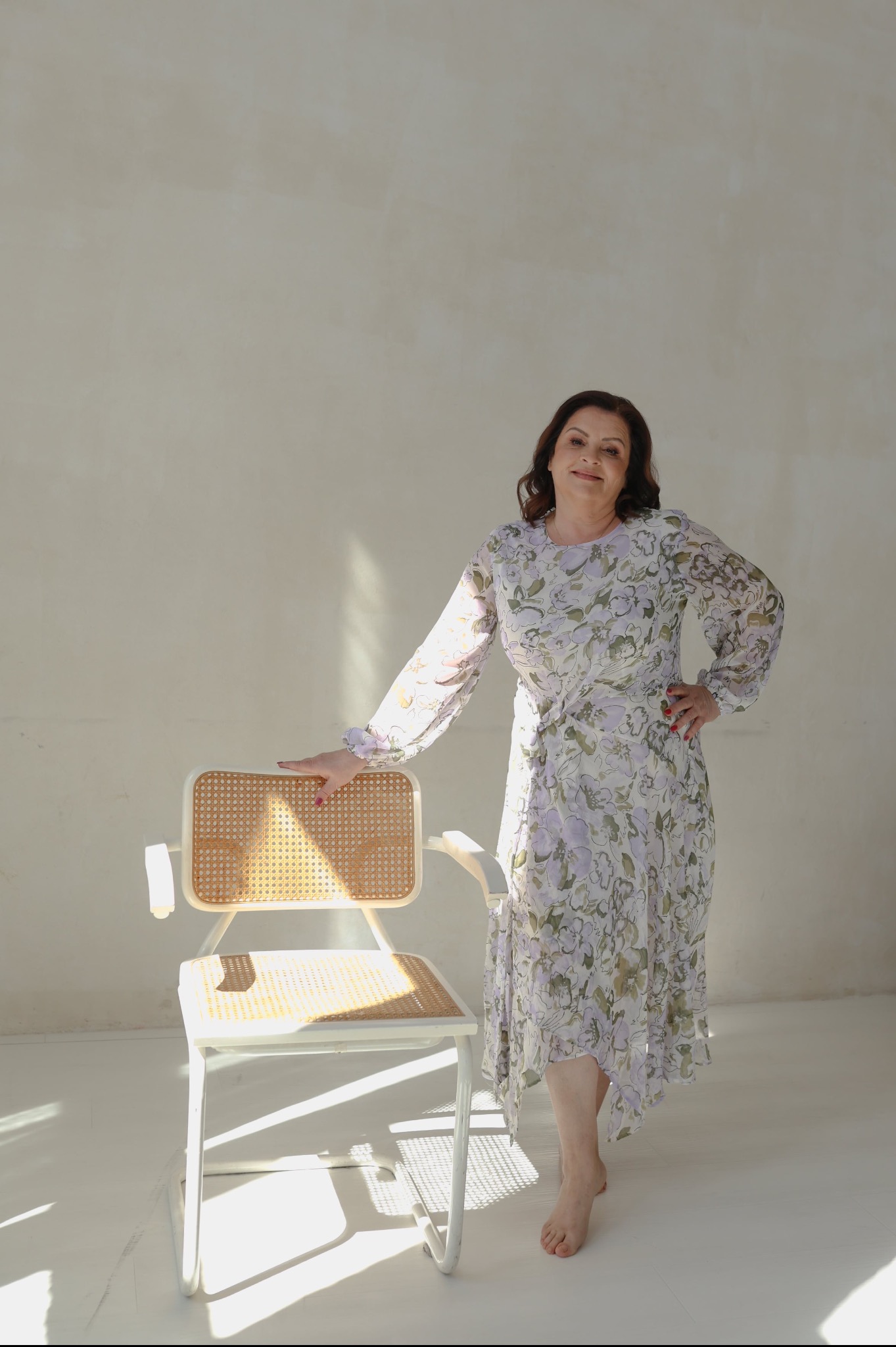 Kobieta w kwiecistej sukience pozuje opierając się o krzesło z rattanowym oparciem w jasnym studio.