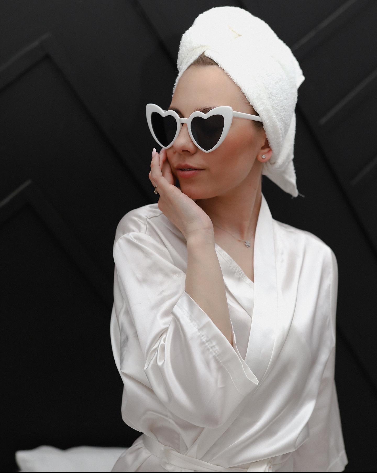 Kobieta w białym szlafroku i ręczniku na głowie, pozująca w okularach w kształcie serc na tle czarnej ściany z geometrycznym wzorem.