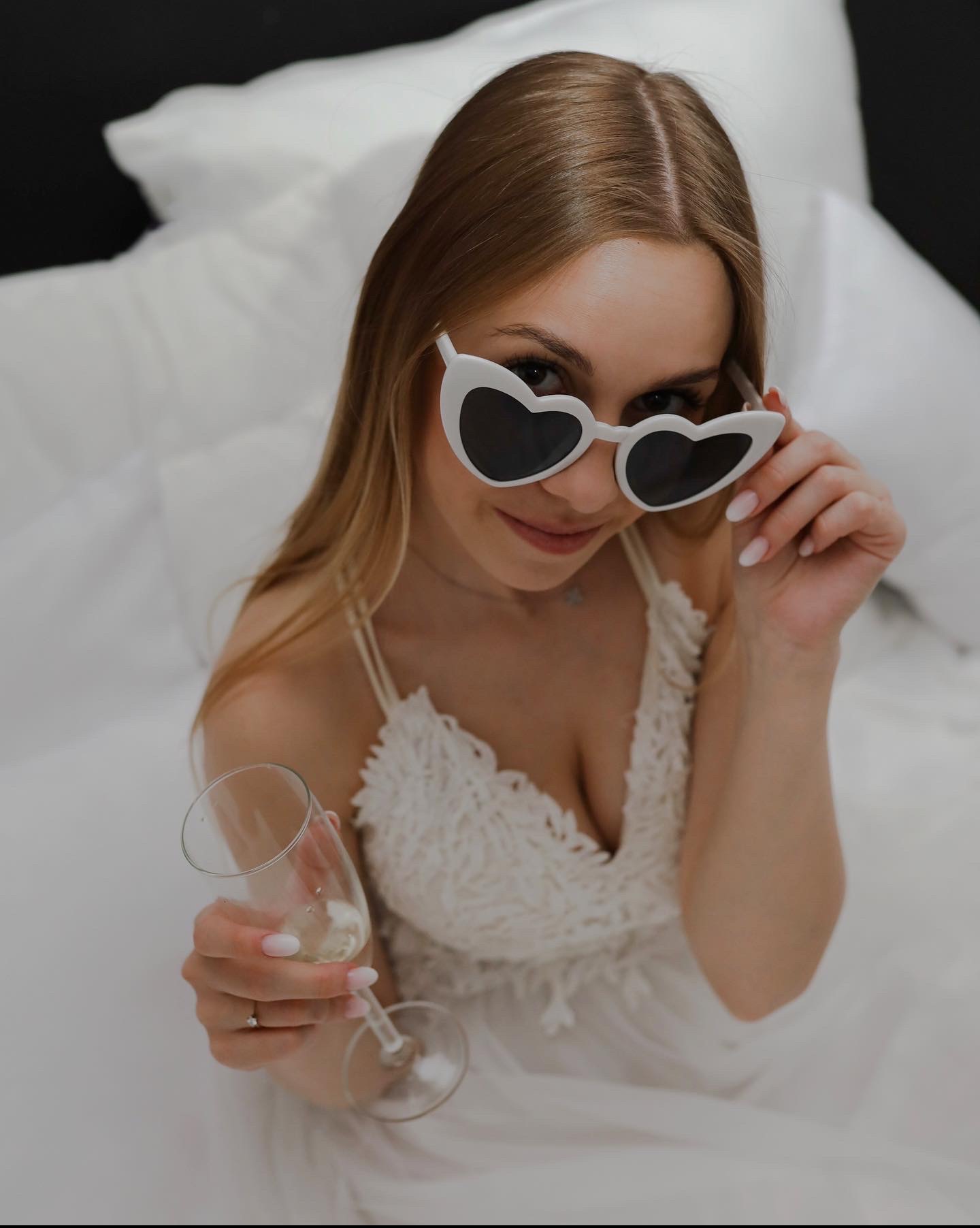 Uśmiechnięta panna młoda w białej sukni, trzymająca kieliszek szampana i okulary w kształcie serc, leżąca na białej pościeli.