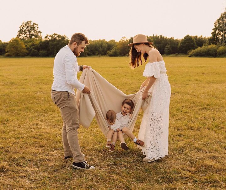 Rodzina z dwójką dzieci na łące, rodzice trzymają koc tworząc hamak dla dzieci, ciepłe światło zachodzącego słońca.