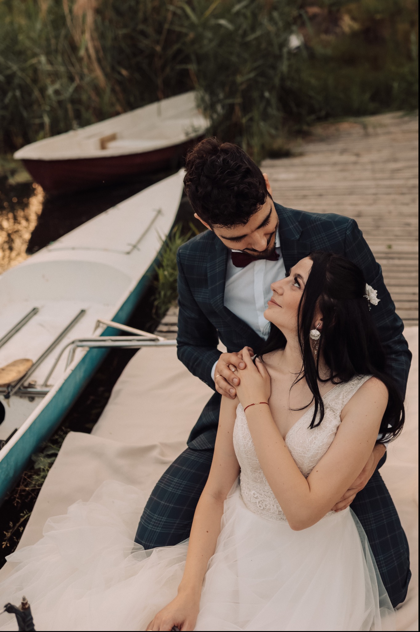 Para młoda w eleganckich strojach pozuje na pomoście obok łodzi, panna młoda w koronkowej sukni z tiulowym dołem spogląda na pana młodego w kraciastej marynarce i bordowej muszce.
