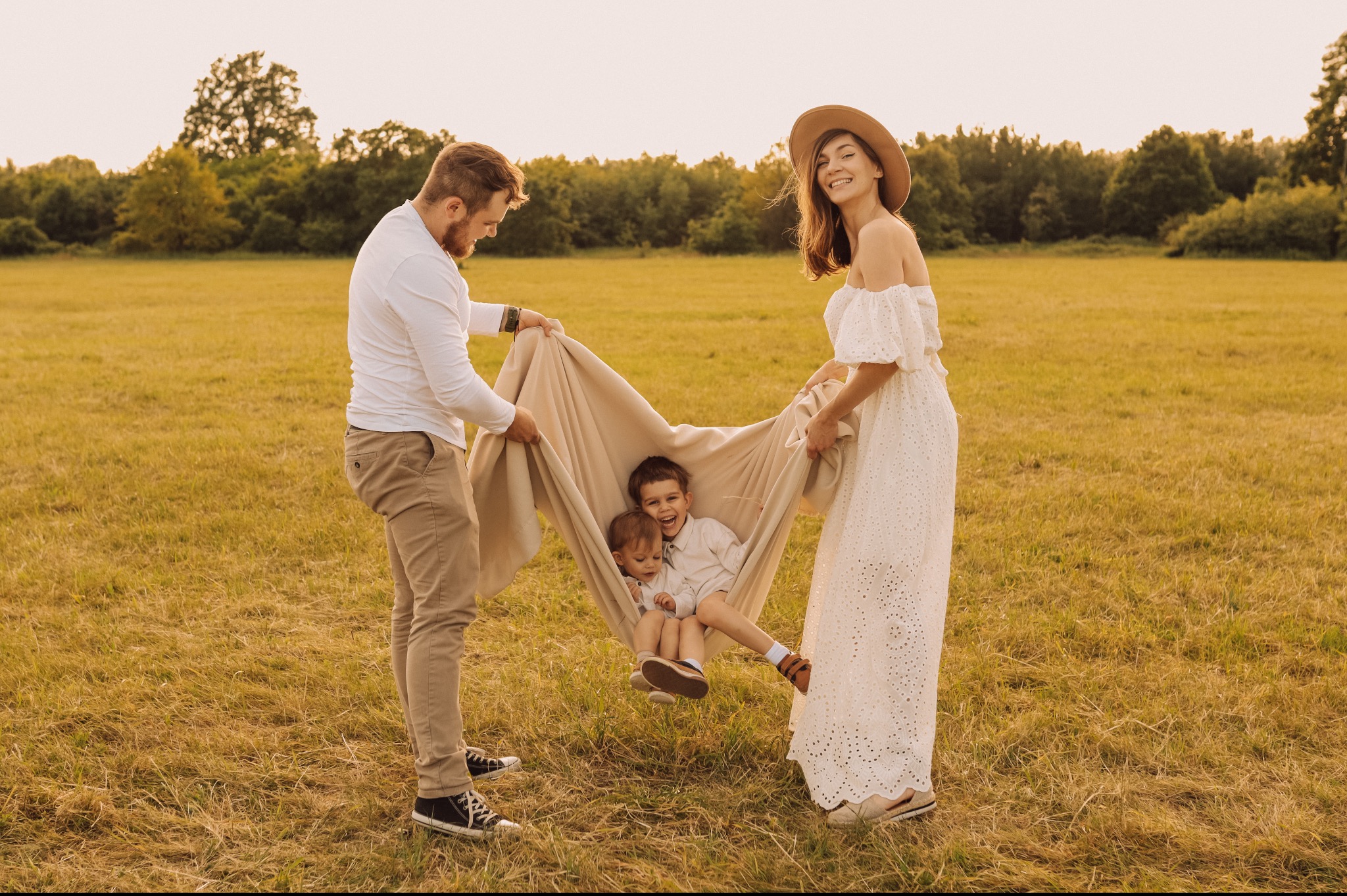 Rodzina z dwójką dzieci bawi się na łące, trzymając koc jak hamak, tata w trampkach, mama w ażurowej sukience i kapeluszu, ciepłe światło zachodzącego słońca.
