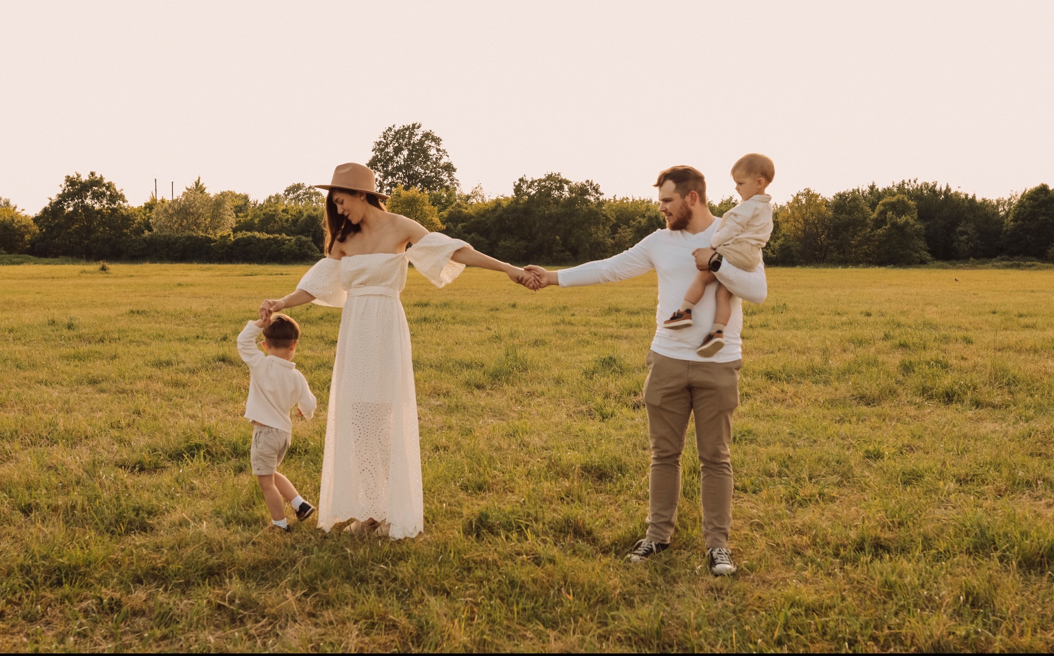 Sesja rodzinna we Wrocławiu: Mama w białej sukience i kapeluszu prowadzi za rękę synka, tata trzyma drugiego synka na rękach, wszyscy na łące w ciepłym świetle.