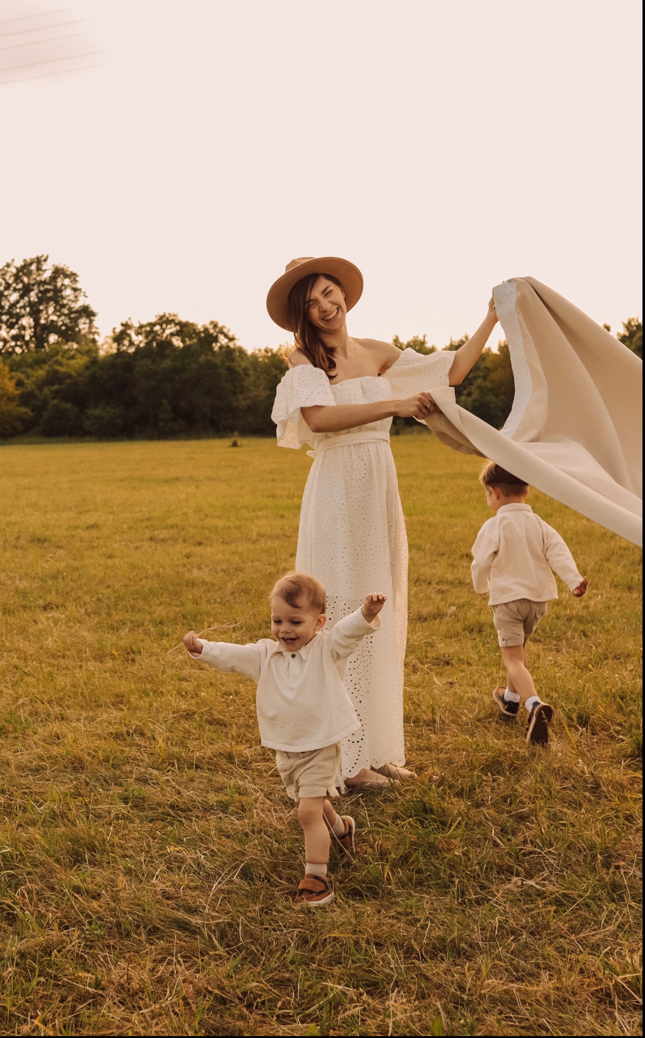 Uśmiechnięta kobieta w białej sukience i kapeluszu trzyma beżowy materiał na łące, obok niej stoi i biegnie dwójka dzieci w jasnych ubraniach.