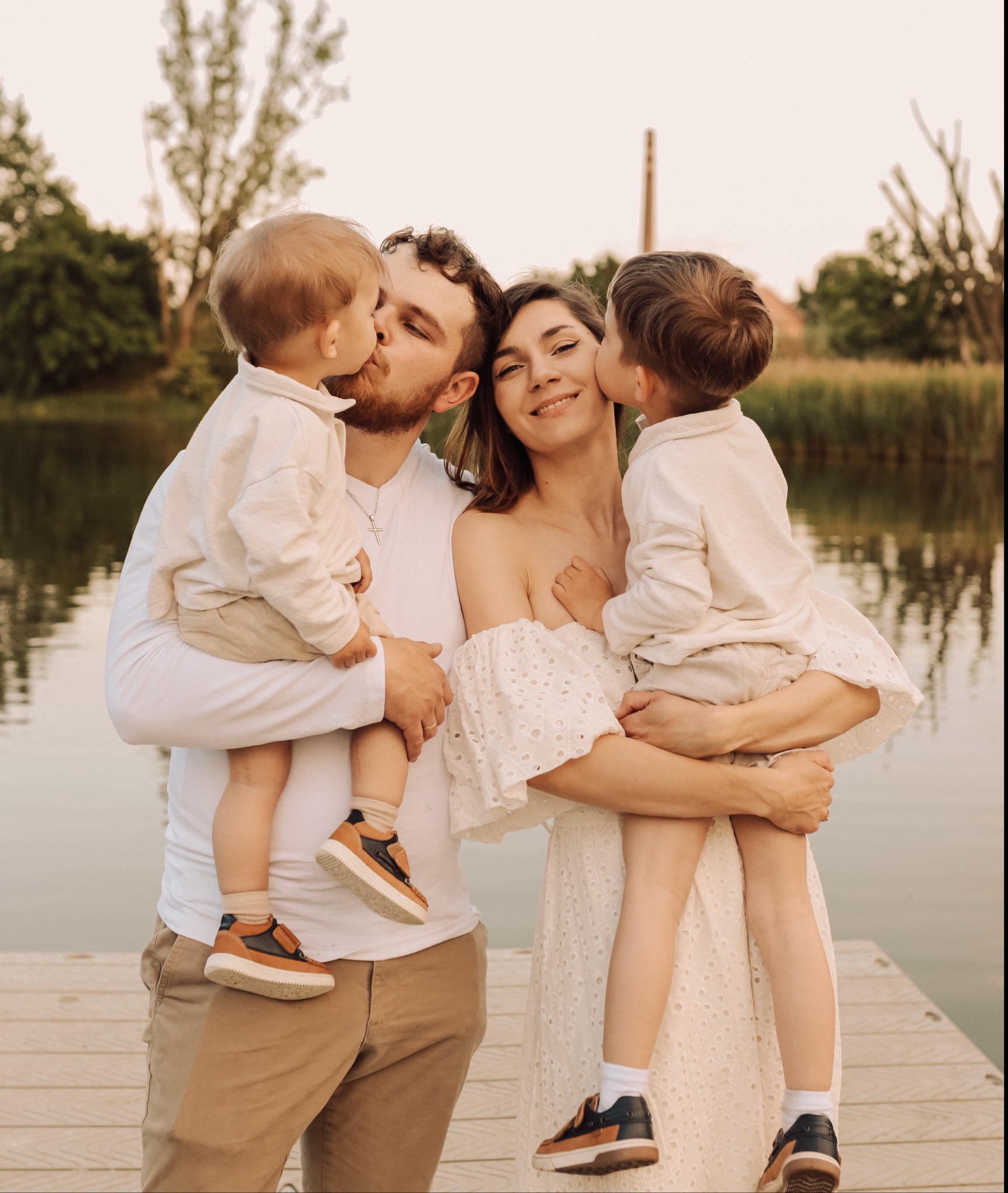 Szczęśliwa rodzina: mama w białej sukience, tata i dwóch synów na drewnianym pomoście nad jeziorem, dzieci całują rodziców w policzki.