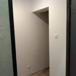 Pomalowane na jasny beż pokoje z widocznymi panelami podłogowymi i czarną futryną drzwiową, widoczny włącznik światła i gniazdko elektryczne.