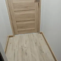 Jasne panele podłogowe ułożone wzdłuż ściany z listwami przypodłogowymi, widoczna drewniana drzwi z poziomymi frezowaniami.