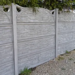 Szary, prefabrykowany płot z betonu imitującego kamień, z dekoracyjnymi wycięciami w górnej części, otoczony żwirem i zielenią, z iglastym drzewem w tle.