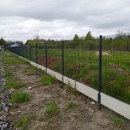 Długi, ciemny panelowy płot z podmurówką z jasnego betonu na nierównym terenie z widokiem na pole i drzewa w tle.