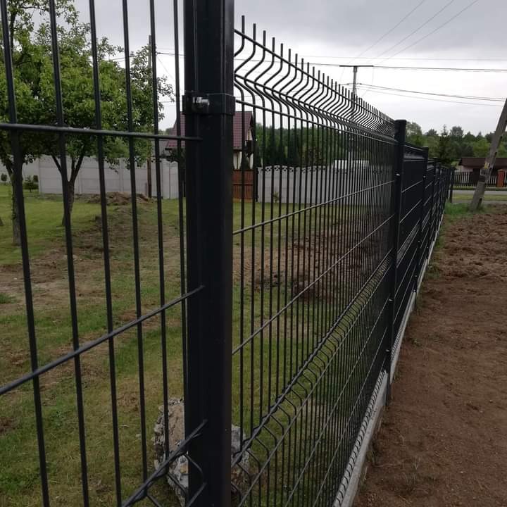 Nowoczesne, czarne ogrodzenie panelowe z widocznymi słupkami i betonową podmurówką, na tle zielonego trawnika i pochmurnego nieba.